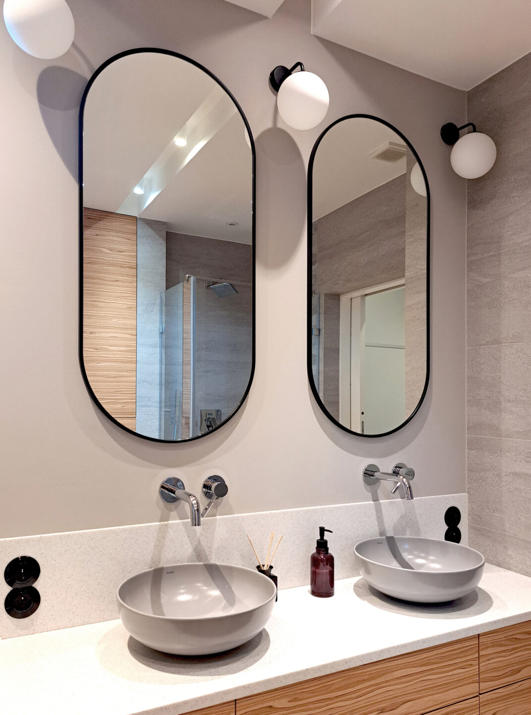 dwa owalne lustra w czarnej ramie nad szarymi umywalkami
