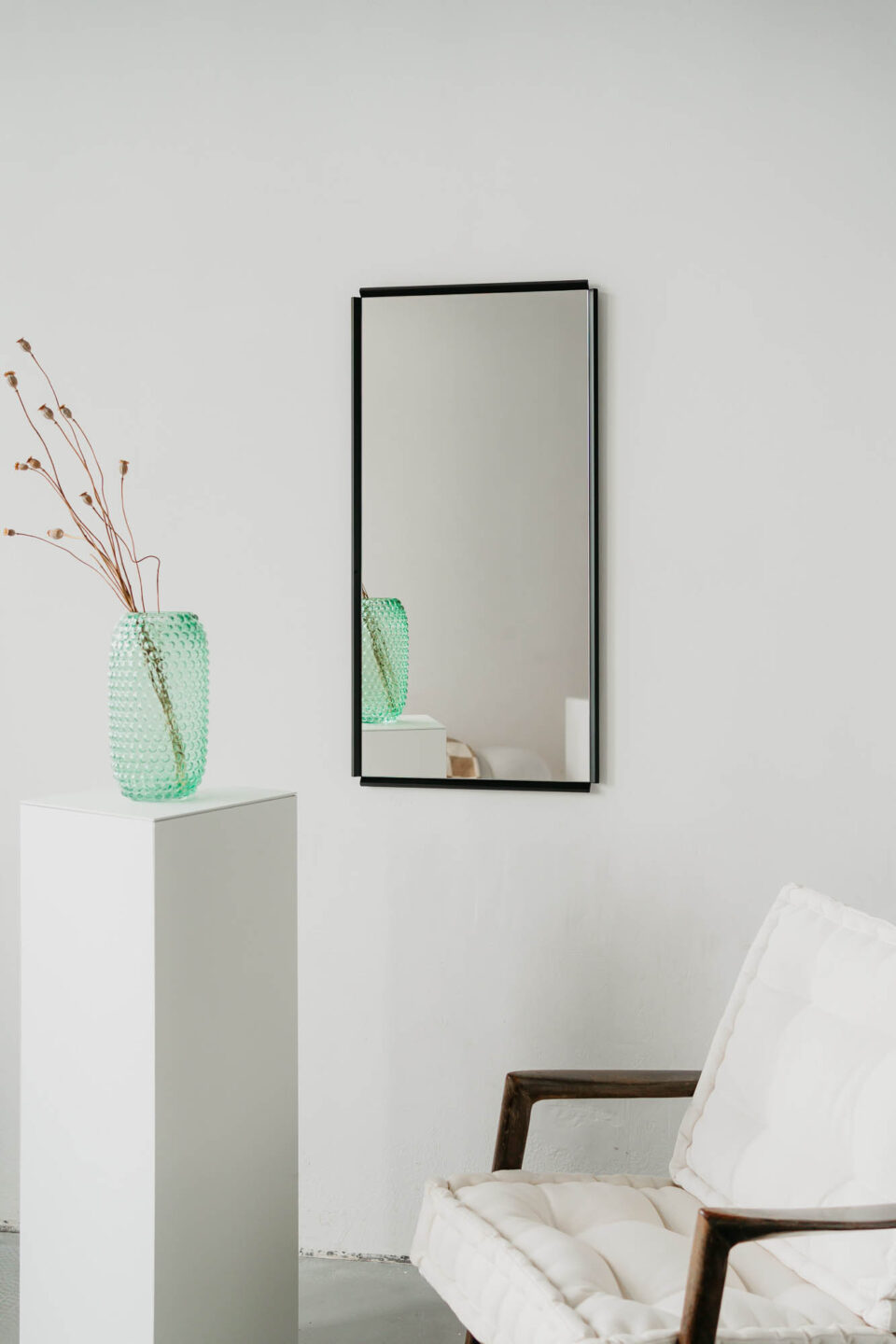 lustro oblique z wycietymi naroznikami. w czarnej ramie na bialej scianie obok bialy fotel i bialy kubik